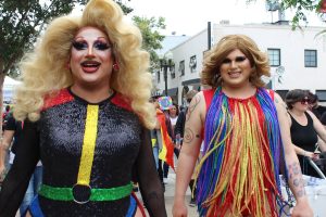 Two white drag queens in blade wigs and colorful attire marching in a Pride parade.