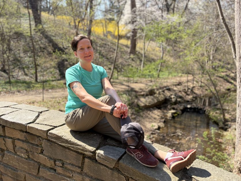Lindsey sitting on a stone wall dressed in a light green shirt and khaki pants. 